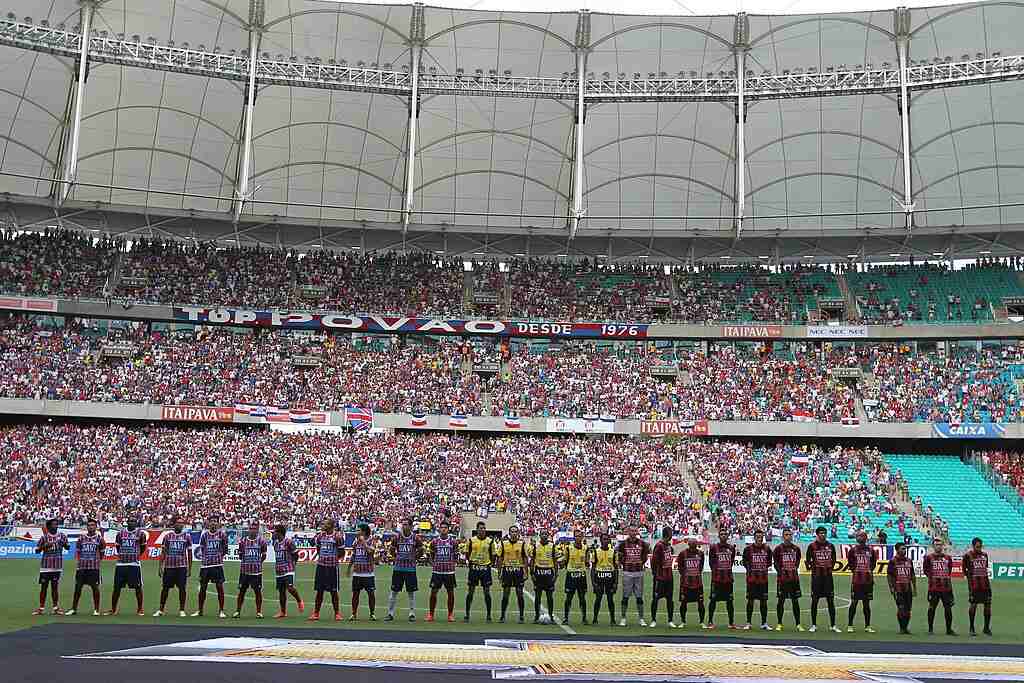 Dois times de futebol enfileirados junto da equipe de arbitragem, em estádio loado, antes do início de uma partida.