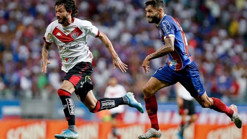 Jogador do Vitória, com uniforme branco e vermelho, conduzindo a bola enquanto jogador do Bahia tenta desarmá-lo.