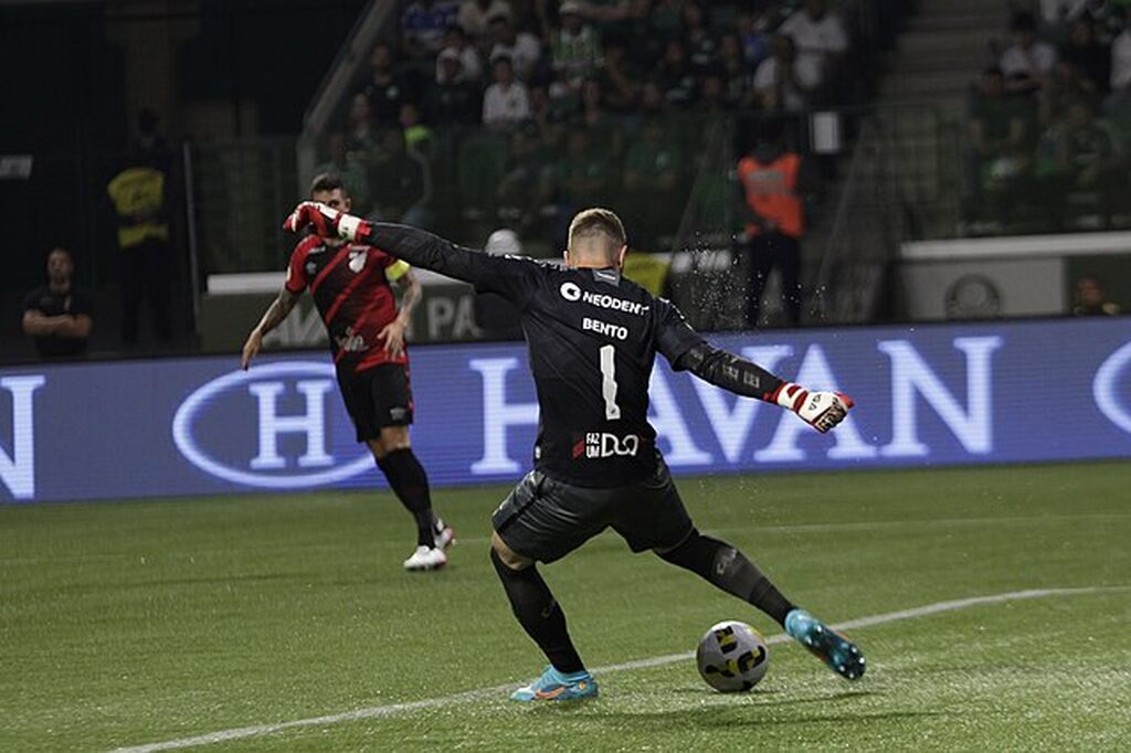 Goleiro de futebol, vestindo uniforme inteiramente preto, chutando a bola durante partida.