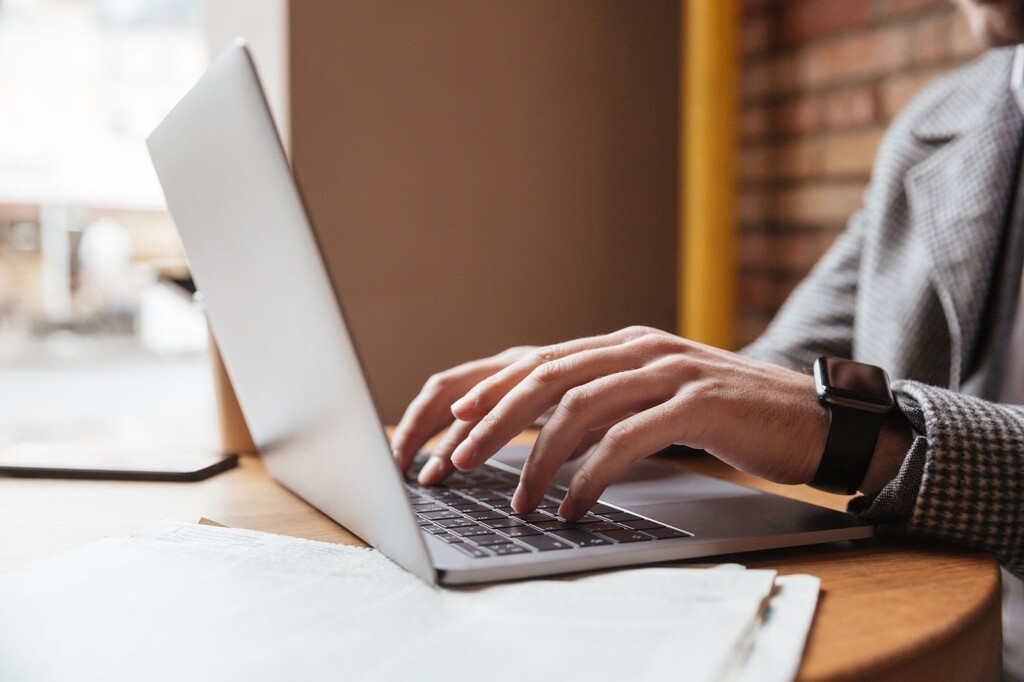 Homem de roupa social digitando em notebook branco sobre uma mesa de madeira com alguns documentos.