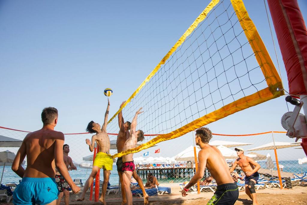 Partida de vôlei sendo disputada na praia, com jogadores tentando bloquear a cortada de jogador adversário.