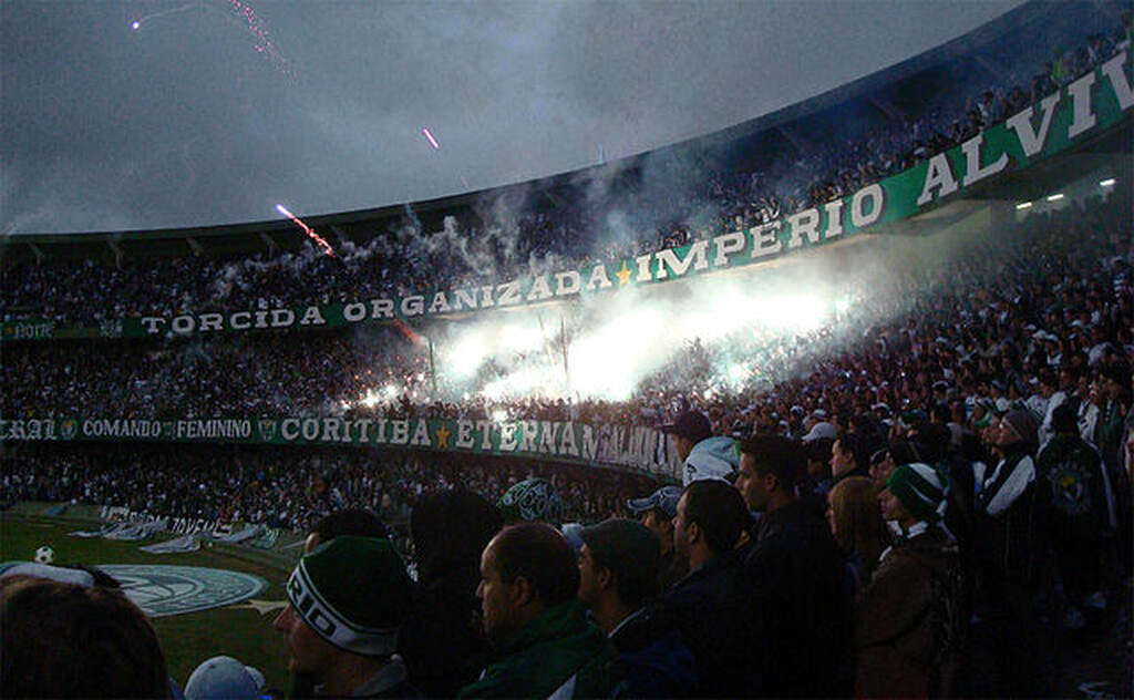 Torcida do Coritiba lotando estádio, soltando fogos e festejando durante partida de futebol.