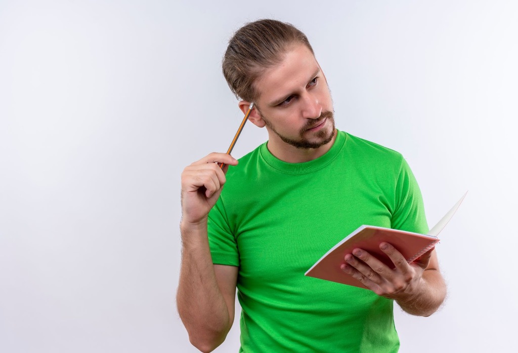 Homem de camisa verde clara, cutucando a cabeça com um lápis enquanto segura um caderno rosa com a mão esquerda.