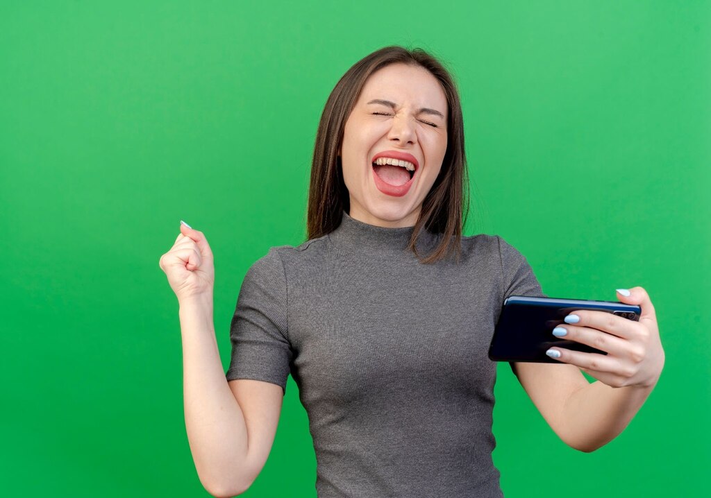 Mulher de roupa cinza, segurando celular com a mão esquerda, enquanto comemora intensamente em um fundo verde.