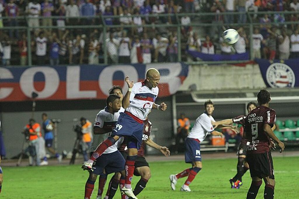 Jogador do Bahia, com uniforme branco, vermelho e azul, saltando para cabecear a bola durante partida.