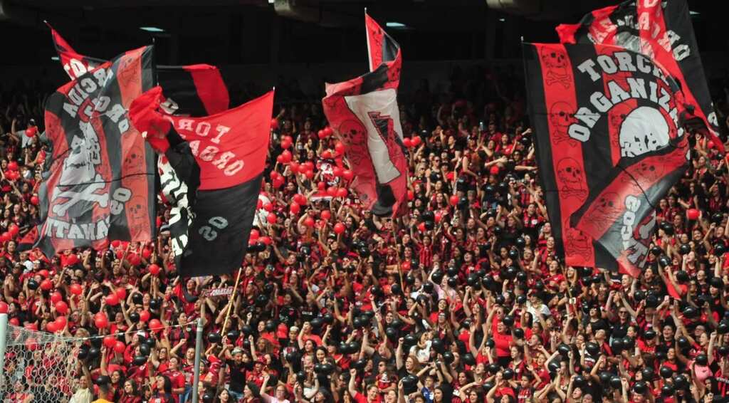 Torcida do Athlético PR, estida com uniforme vermelho e preto do clube, fazendo festa com bandeiras no estádio.