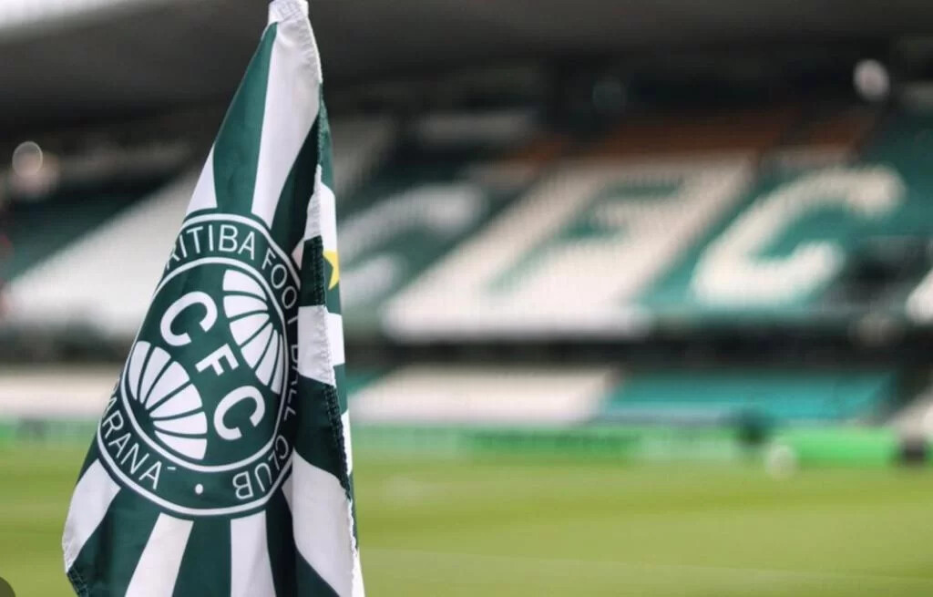 Haste de bandeira de escanteio com a bandeira do Coritiba pendurada, com vista para arquibancada do estádio ao fundo.