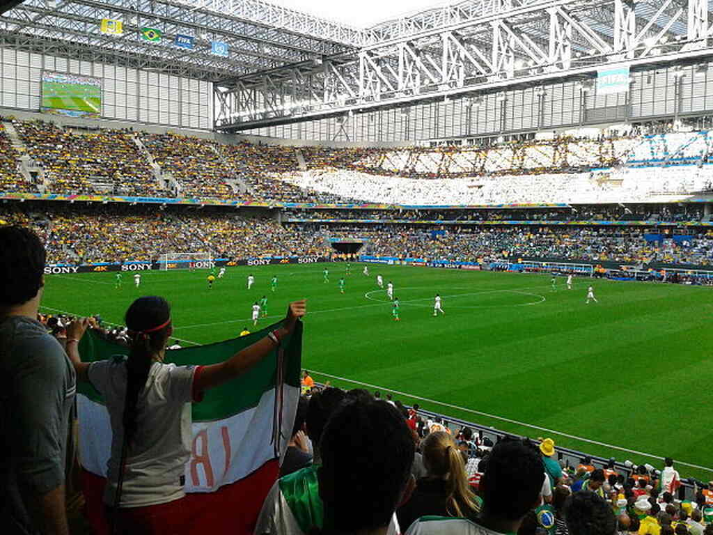 Estádio de futebol lotado, com vista da arquibancada para o campo durante partida.