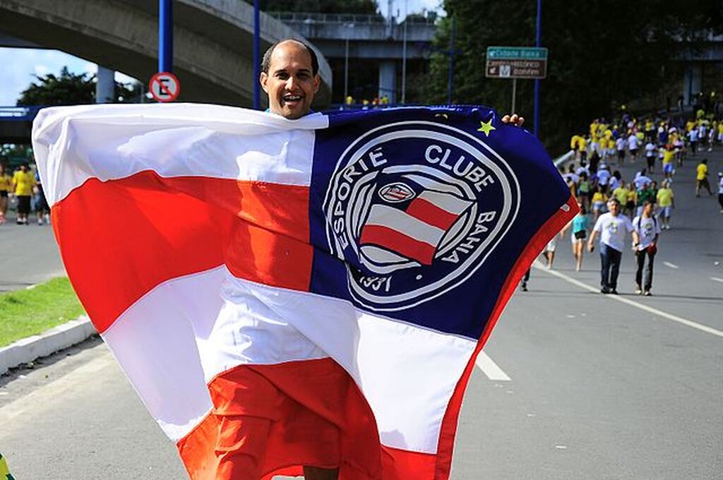 Homem segurando bandeira do Bahia no meio da rua, com uma multidão de pessoas andando atrás dele.