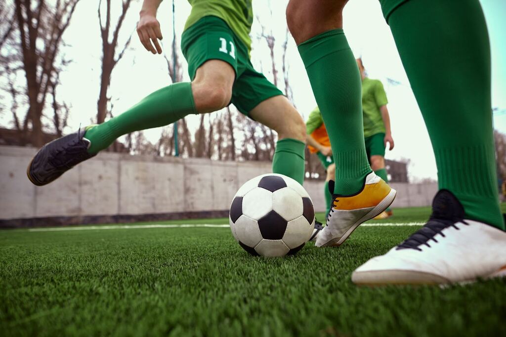 Dois jogadores de futebol vestindo uniformes e meiões verdes, dividindo uma bola durante partida de futebol no gramado.