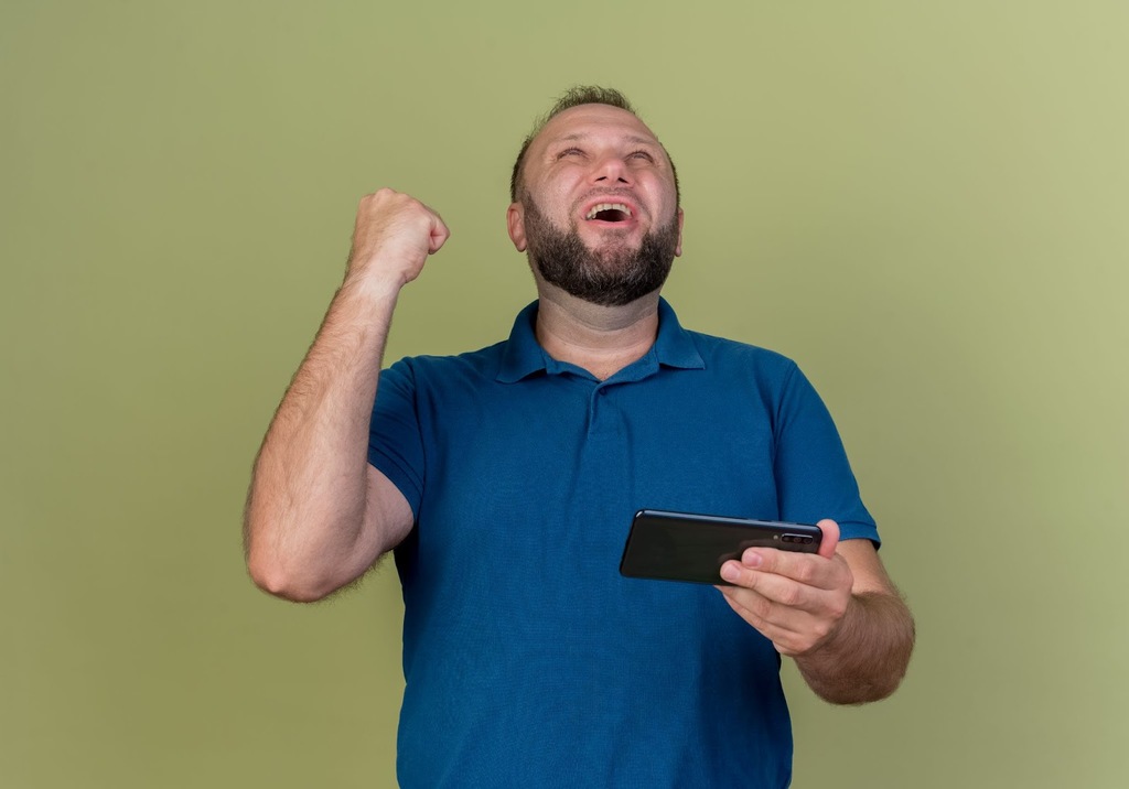 Homem de camisa azul segurando um celular com a mão esquerda, olhando para o alto e comemorando.