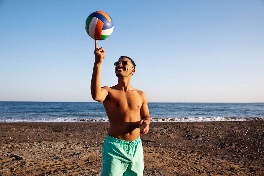 Homem de calção verde claro, com uma bola de vôlei equilibrada na ponta de seu dedo, na praia, perto do mar.