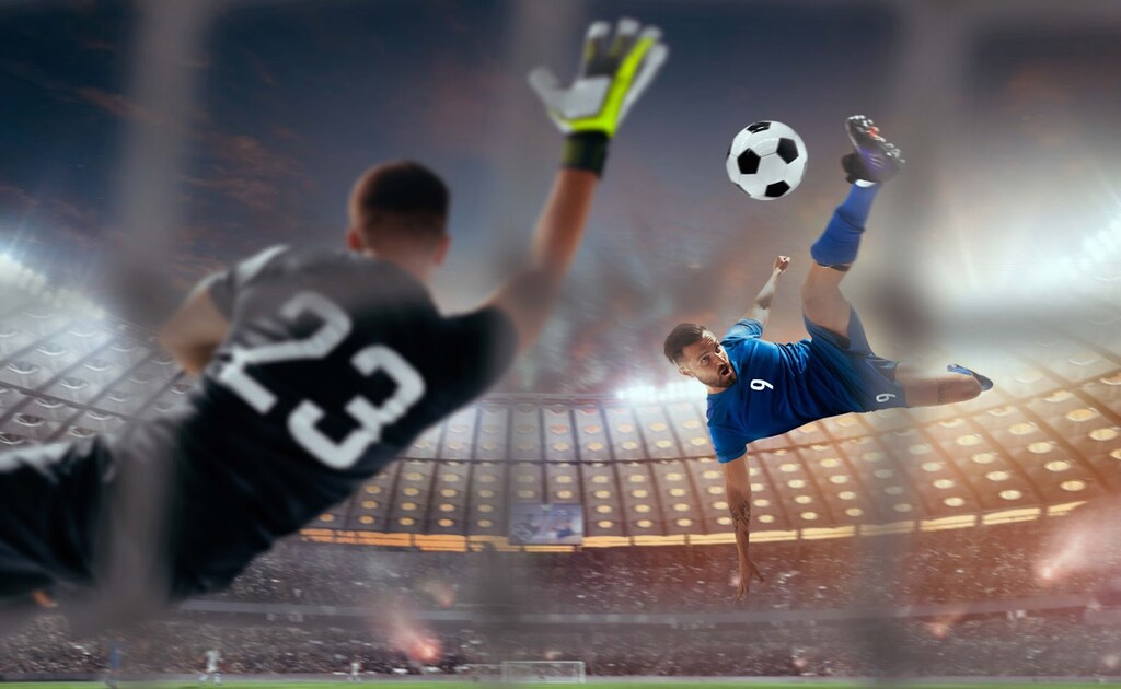 Jogador de futebol de uniforme azul, dando um voleio na bola, enquanto o goleiro tenta defender.