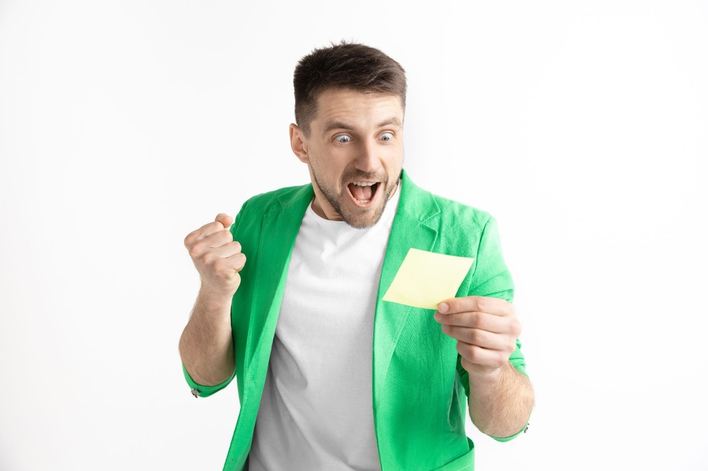 Homem de camisa branca vestindo um casaco verde e comemorando enquanto olha para um bilhete em sua mão esquerda.