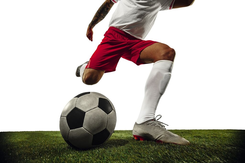 Jogador de futebol de camiseta branca e calção vermelho, no gramado, executando o movimento para chutar a bola de futebol.