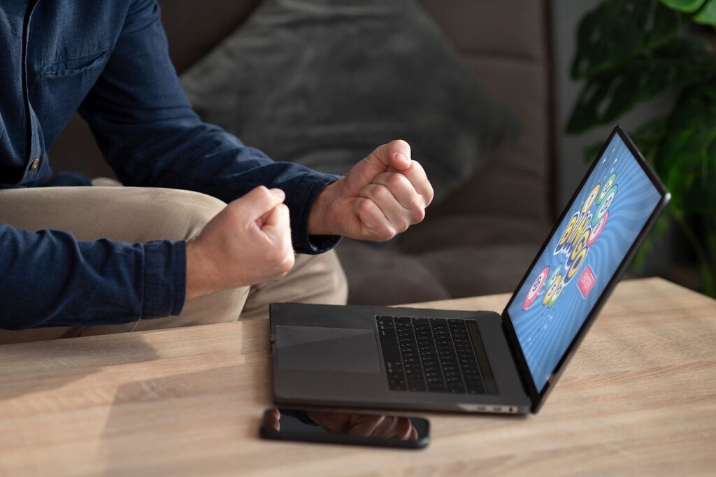 Pessoa de blusa de manga comprida, com os punhos fechados, comemorando, em frente a um notebook preto sobre a mesa de madeira