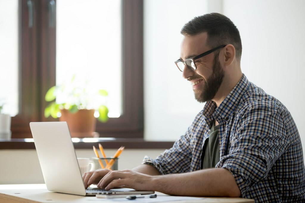 Homem de camisa social, barba e óculos digitando em um notebook branco sobre a mesa.