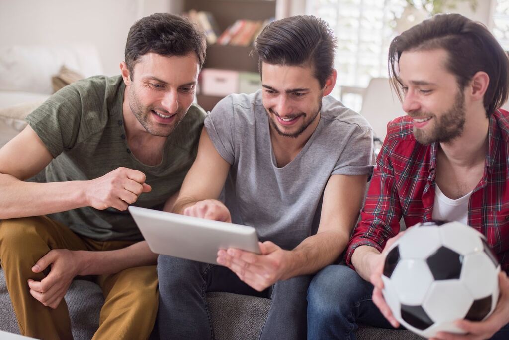 3 homens sentados em um sofá, olhando para um tablet e segurando uma bola de futebol enquanto sorriem.