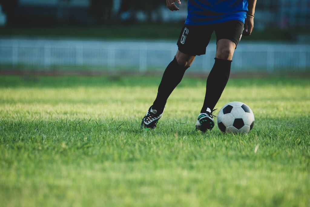 Jogador de futebol de calção preto e camisa azul escura, conduzindo uma bola de futebol pelo gramado.