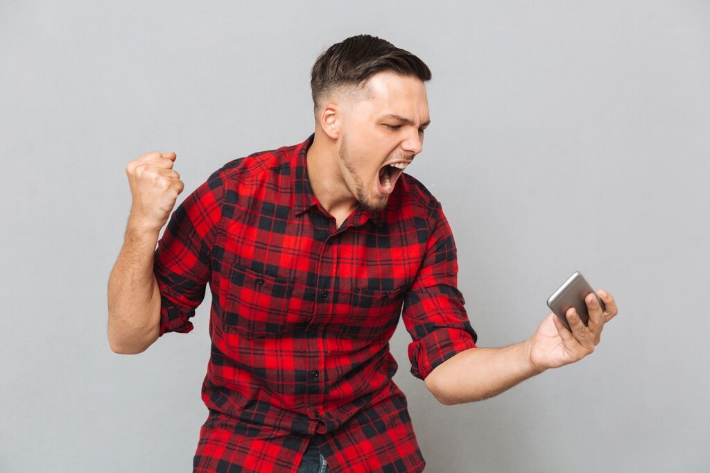 Homem de camisa xadrez vermelha e preta, comemorando enquanto segura e olha para o smartphone, diante de um fundo cinza.