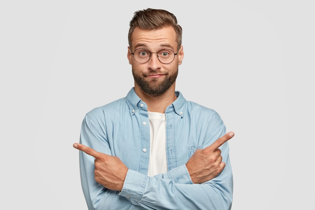 Homem de óculos, camisa social azul aberta, com os braços cruzados e apontando os dedos indicadores para direções opostas.