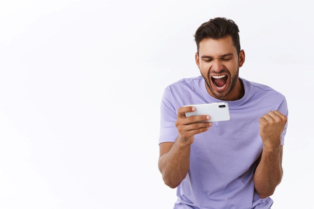 Homem de camiseta lilás, comemorando com expressão feliz, enquanto segura um smartphone branco, diante de um fundo branco.