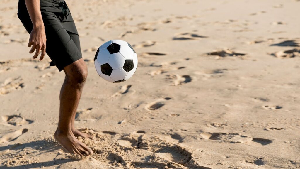 Homem com calção preto fazendo embaixadinhas com bola de futebol na areia da praia.