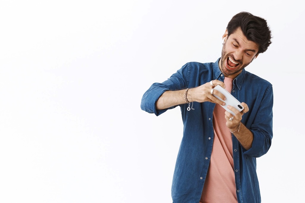 Homem de camisa jeans aberta por cima de uma camiseta rosa, comemorando enquanto olha para o smartphone.