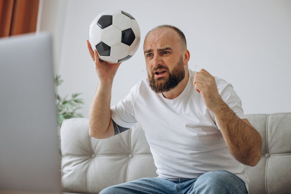 Homem nervoso, de camiseta branca e calça jeans, sentado no sofá olhando para frente, enquanto segura uma bola.