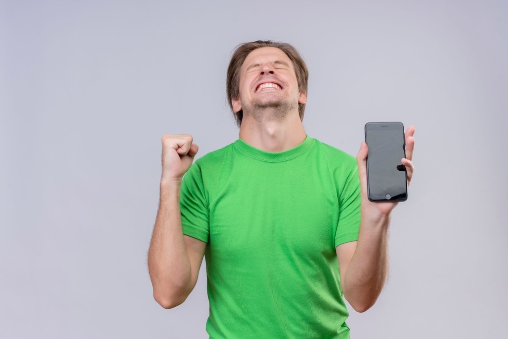 Homem de camiseta verde, comemorando enquanto segura o smartphone, diante de um fundo cinza.