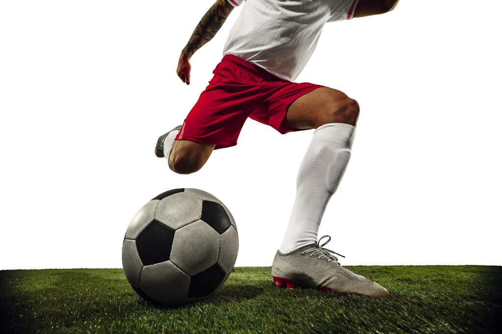 Jogador de camiseta branca e calção vermelho, no gramado fazendo o movimento para chutar a bola de futebol.