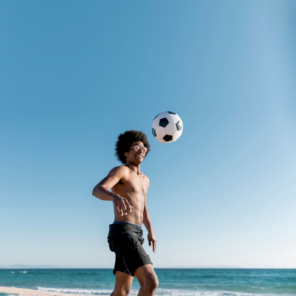 Homem sem camisa e de bermuda preta, na praia, observando a bola de futebol no alto acima da sua cabeça.