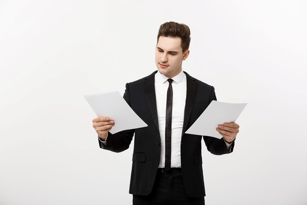 Homem de terno e gravata pretos e camisa social branca, segurando e olhando para duas folhas, na frente de um fundo branco.