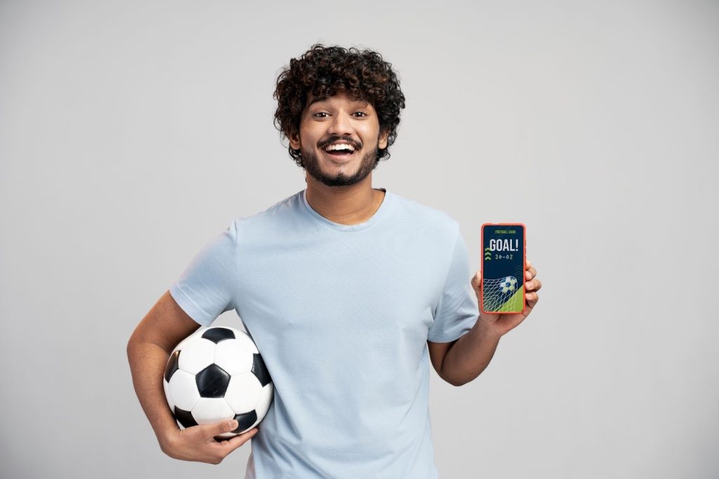 Homem de camiseta azul claro, sorrindo enquanto segura uma bola de futebol e mostra um smartphone com um alerta de gol.