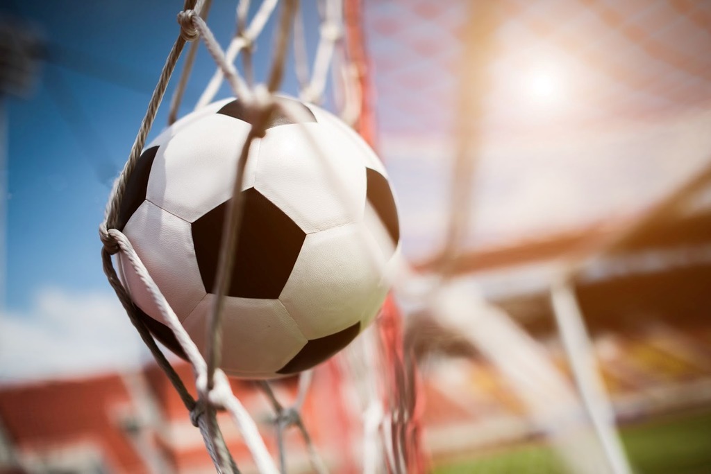 Bola de futebol clássica com pentágonos pretos e hexágonos brancos, estufando as redes, com o sol brilhando ao fundo.