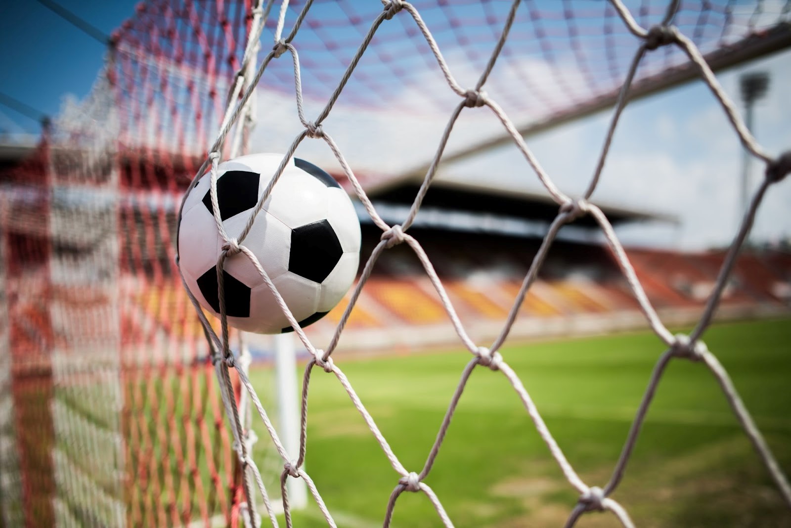 Uma bola de futebol batendo e estufando as redes de um gol em um estádio de futebol com as arquibancadas vazias.