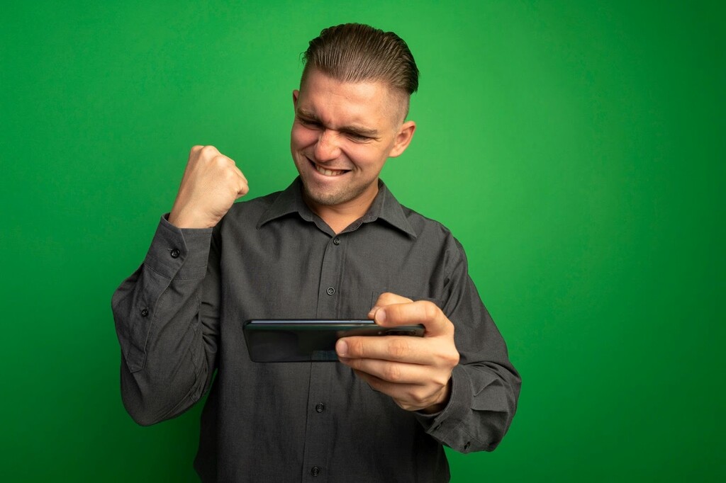 Homem de camisa cinza escuro, comemorando enquanto segura e olha para o smartphone, diante de um fundo verde.