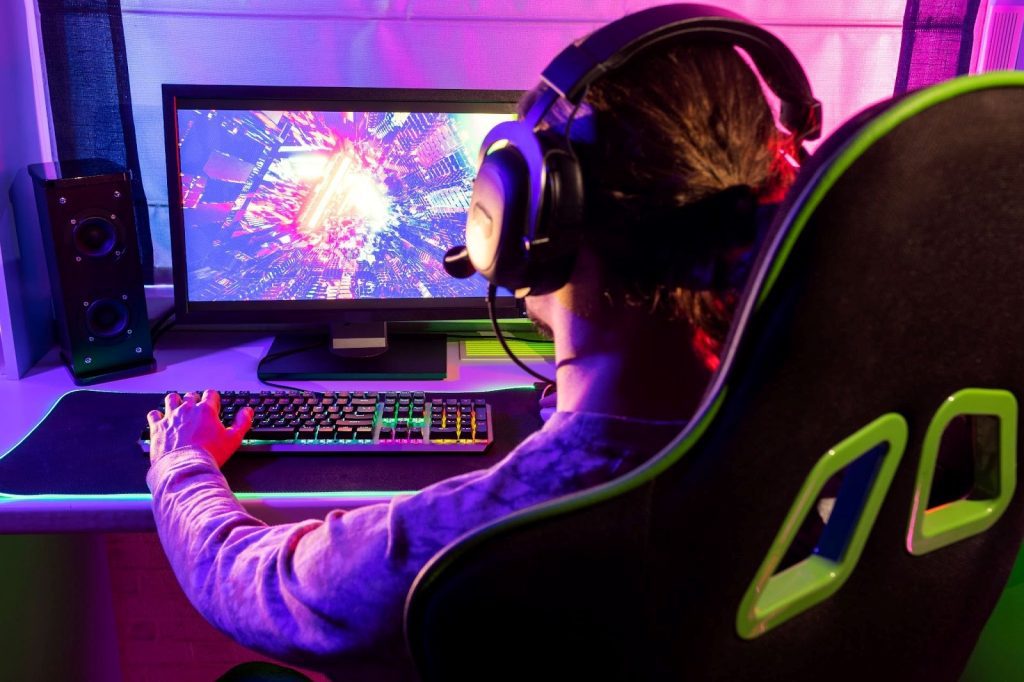 Homem sentado em uma cadeira gamer, na frente de uma mesa com um monitor com a tela cheia de luzes, mexendo no teclado.