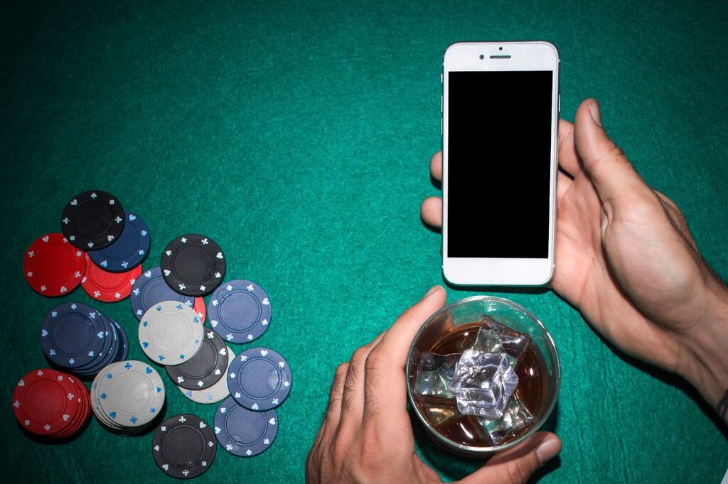 Homem segurando um smartphone e um copo de bebida com gelo, apoiado em uma mesa de poker com algumas fichas ao seu lado.