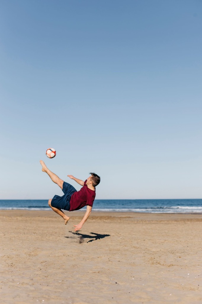 Homem de camiseta vinho e calção azul marinho, na praia, fazendo movimento de bicicleta para acertar a bola.