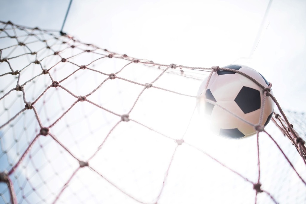 Bola de futebol clássica com pentágonos pretos e hexágonos brancos, estufando as redes, com o sol brilhando ao fundo.