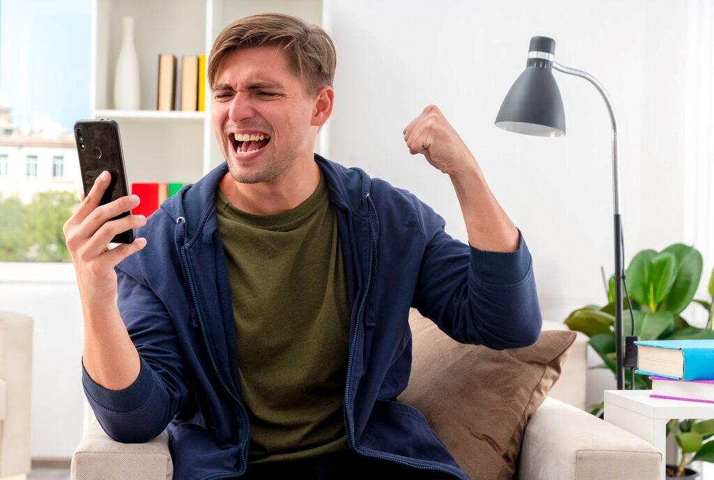 Homem de camiseta verde e jaqueta azul, sentado no sofá, sorrindo e comemorando, enquanto segura e olha para o smartphone.