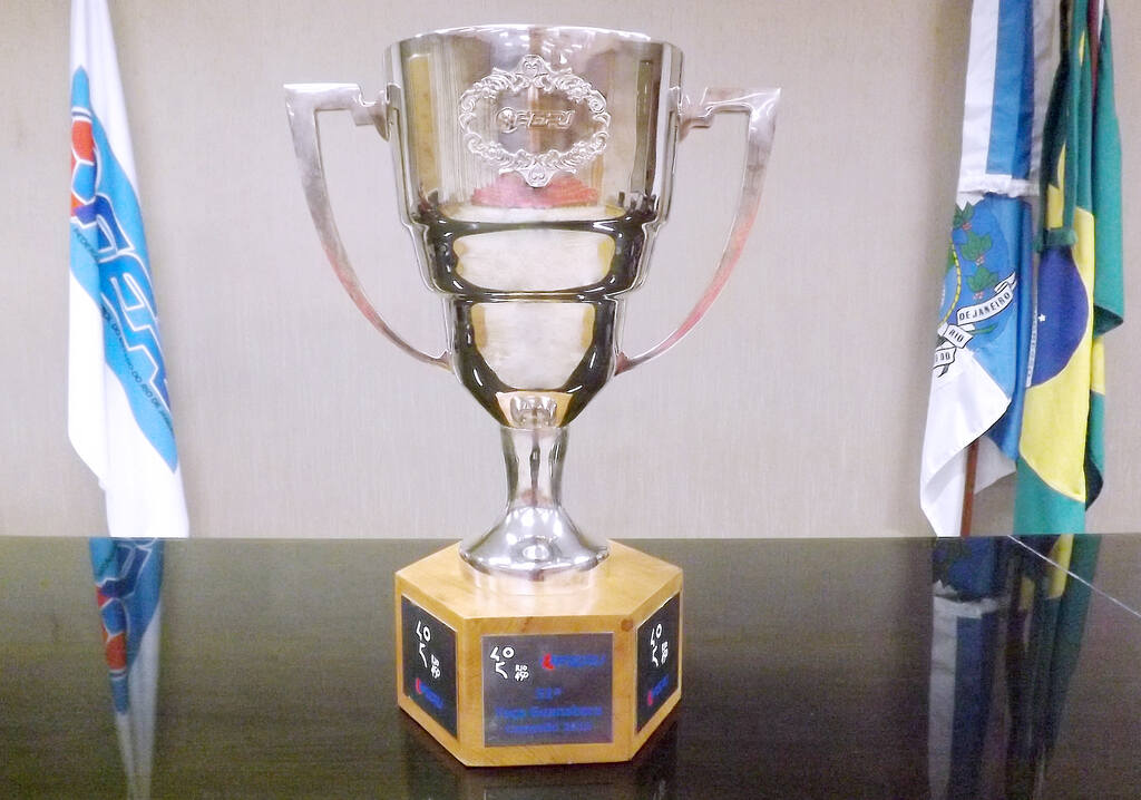 Taça Guanabara em cima de uma mesa com as bandeiras do Brasil, do estado e da cidade do Rio de Janeiro atrás.