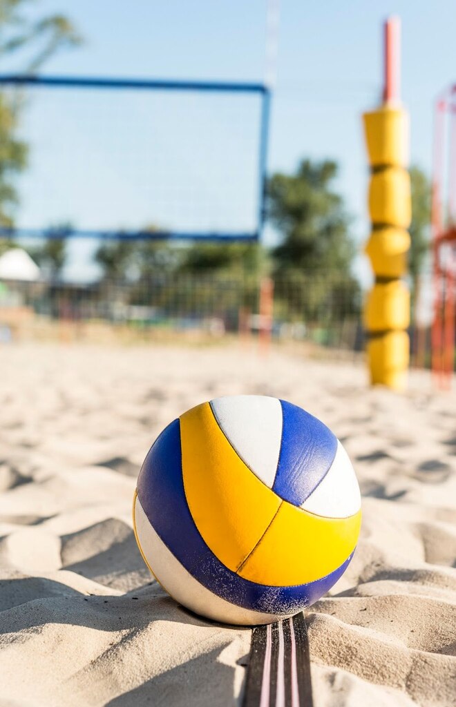 Bola de vôlei na areia em uma quadra de vôlei de praia, com a rede desfocada ao fundo.