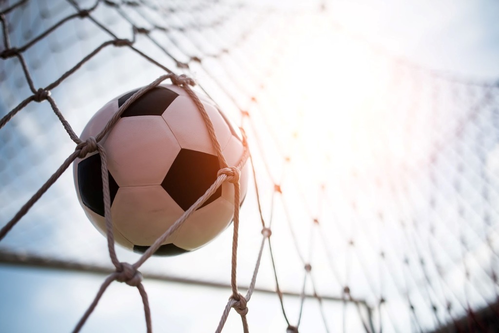 Bola de futebol clássica com pentágonos pretos e hexágonos brancos, estufando as redes, com o sol brilhando ao fundo.