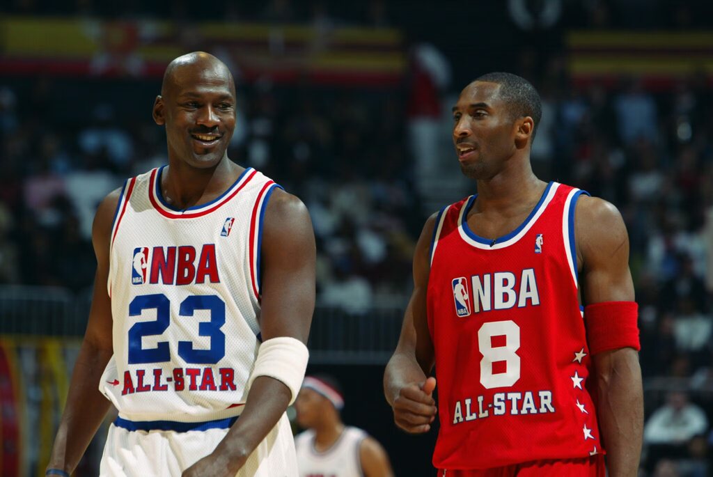 Michael Jordan de uniforme branco durante o All Star Game NBA ao lado de jogador de uniforme vermelho andando pela quadra.