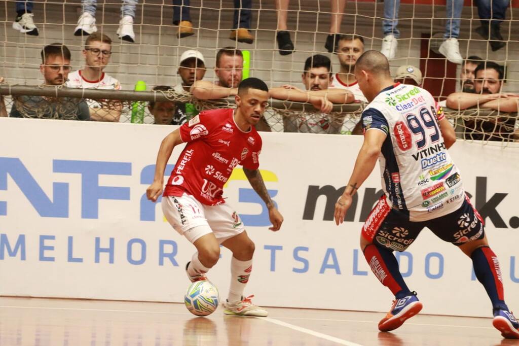 Jogador de futsal vestindo uniforme vermelho e calção branco tentando drible em jogador de uniforme azul e branco.