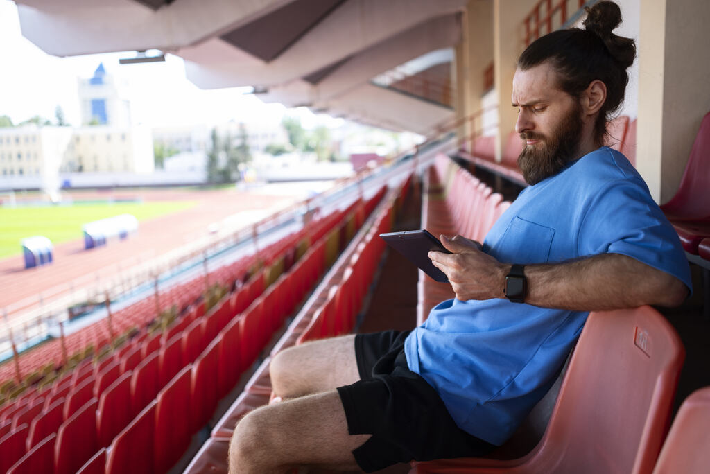 Homem de camisa azul, coque samurai e barba, usando um tablet sentado em cadeiras vermelhas em uma arquibancada.