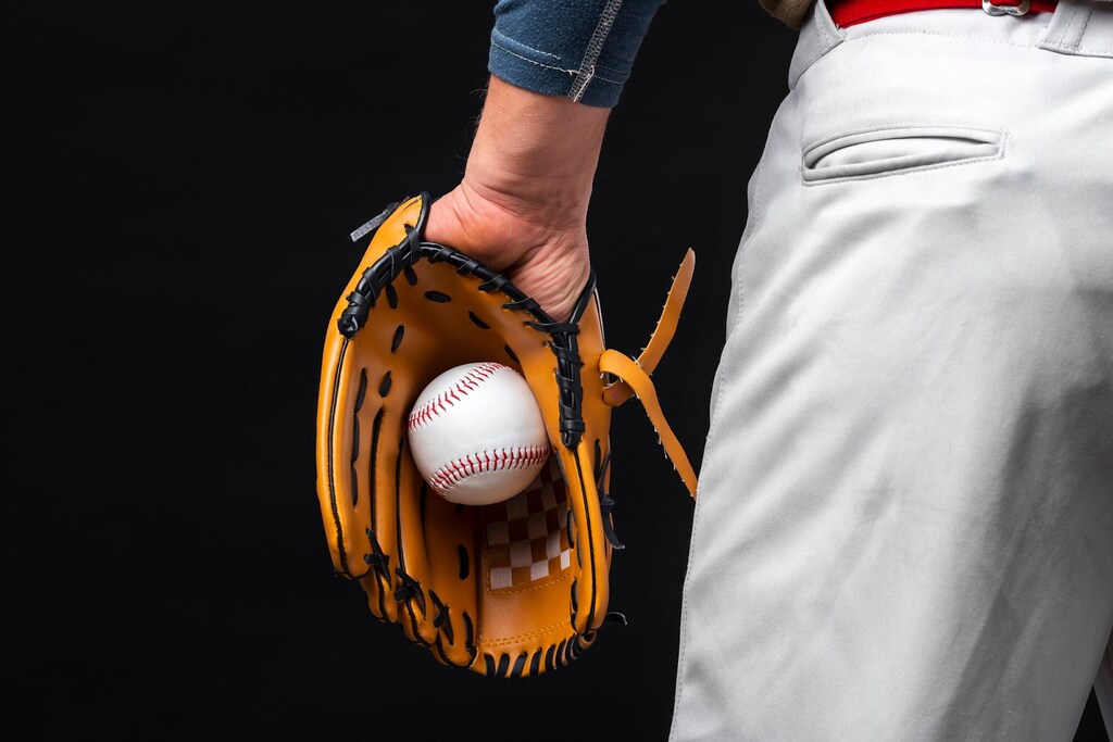 Jogador de baseball visto de costa, segurando uma bola de baseball, com luva marrom, na mão esquerda.