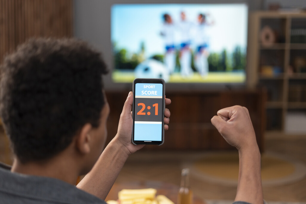 Homem acompanhando um placar pelo celular enquanto assiste um jogo de futebol na TV e comemora.